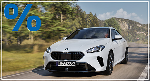 Das Bild zeigt einen weißen, viertürigen BMW 2er-Reihe Gran Coupé, der auf einer kurvigen Straße fährt. Das Auto ist schräg von vorne zu sehen. Im Vordergrund ist ein großes, rotes Prozentzeichen (%) platziert. Der Hintergrund zeigt einen Wald und Berge unter einem blauen Himmel. Das Kennzeichen "M ZJ 6656" ist auf der vorderen Stoßstange zu sehen.