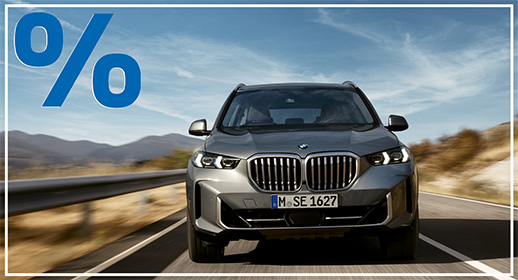Das Bild zeigt ein graues SUV, den BMW X5, der auf einer Landstraße fährt. Das Auto ist von vorne zu sehen. Im Vordergrund, über dem Auto, ist ein großes, rotes Prozentzeichen (%) platziert. Der Hintergrund zeigt eine bergige Landschaft unter einem blauen Himmel mit einigen Wolken.