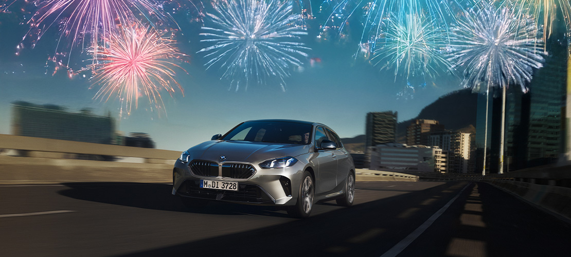 Diese Werbegrafik präsentiert einen silbergrauen BMW 1er (Modell 120) während der Fahrt vor einer unscharfen Stadtkulisse. Wie bei den anderen Aktionsgrafiken ist oben links ein markantes blaues Prozentzeichen (%) zu sehen, und zwei helle Feuerwerke erhellen den Himmel über dem Fahrzeug. Das Bild vermittelt urbane Agilität und weist auf die speziellen Konditionen für dieses Modell bei Rhein BMW hin.