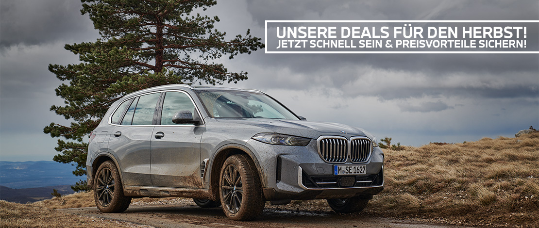 Das Bild zeigt ein graues BMW SUV (wahrscheinlich ein X5), das leicht mit Schmutz bedeckt ist. Das Auto steht auf einem Schotterweg in einer natürlichen, herbstlichen oder bergigen Umgebung. Im Hintergrund sind eine große Kiefer und eine weite Landschaft unter einem bedeckten, dramatischen Himmel zu sehen.  Text im Bild:  Über dem Auto befindet sich ein weißes Banner mit schwarzer Schrift. Der Text lautet:  "UNSERE DEALS FÜR DEN HERBST! JETZT SCHNELL SEIN & PREISVORTEILE SICHERN!"