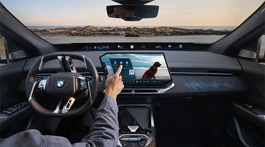 Moderner Innenraum des neuen BMW iX4 mit BMW Lenkrad, großem zentralem Touchdisplay und digitaler Instrumentenanzeige. Eine Person bedient den Touchscreen, während die beleuchteten Designelemente des Armaturenbretts und die moderne Mittelkonsole zu sehen sind.