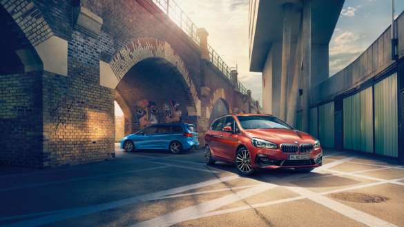 Ein roter BMW 2er Active Tourer steht auf einem sonnigen Platz unter einer gemauerten Brücke mit Bögen. Im Hintergrund, weiter hinten unter einem anderen Bogen, ist ein blauer BMW zu sehen. Die Szene hat eine städtische, leicht industrielle Atmosphäre mit starkem Gegenlicht.