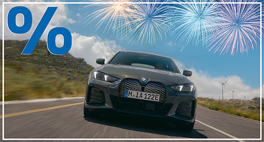 Diese Werbegrafik zeigt einen modernen, dunkelgrauen BMW i4, der dynamisch auf einer Landstraße durch eine hügelige Küstenlandschaft fährt. Die Kamera ist tief positioniert und blickt von vorne auf das vollelektrische Gran Coupé, was die markante Frontpartie und die sportliche Präsenz unterstreicht. Im Hintergrund sind sanfte Hügel, spärliche Vegetation und Windräder unter einem strahlend blauen Himmel mit lockeren Wolken zu sehen.  Der obere rechte Bildbereich wird von mehreren großflächigen, leuchtenden Feuerwerken in Blau- und Goldtönen ausgefüllt. Wie bei allen Motiven dieser Kampagne vervollständigen das große blaue Prozentzeichen oben links und der schmale weiße Außenrahmen das festliche Design zum Jahresstart.