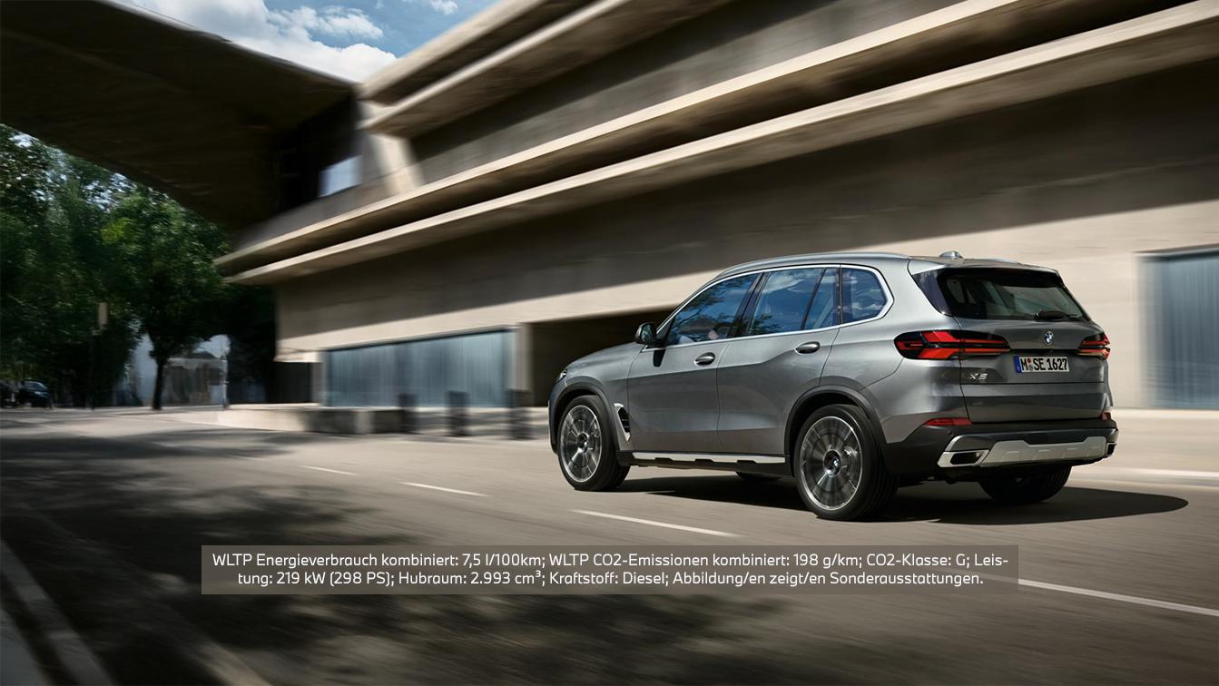 Ein seitlich-hinten aufgenommenes Bild eines grauen BMW X5, der auf einer Straße fährt. Im Hintergrund sind moderne Gebäude zu sehen, die verschwommen wirken, was Bewegung andeutet. Das Fahrzeug hat dynamische Linien und glänzende Felgen.  Über dem Bild ist ein Textoverlay mit folgenden Informationen: WLTP Energieverbrauch kombiniert: 7,5 l/100km; WLTP CO2-Emissionen kombiniert: 198 g/km; CO2-Klasse: G; Leistung: 219 kW (298 PS); Hubraum: 2.993 cm³; Kraftstoff: Diesel; Abbildung/en zeigt/en Sonderausstattungen.