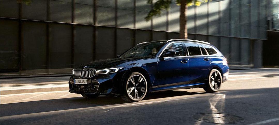 Top Deal BMW 3er Touring Dunkelblaue BMW 3er Touring Kombi in Bewegung auf einer städtischen Straße, mit einem modernen Gebäude und Bäumen im Hintergrund. Das Bild zeigt das spezifische Modell des Angebots dynamisch
