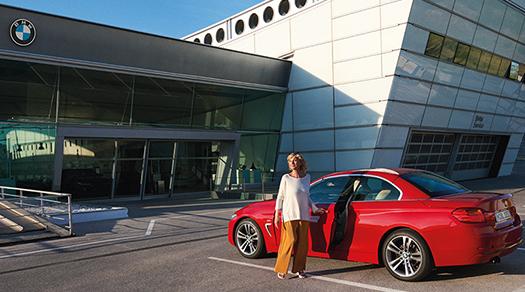 Frau steht lächelnd neben rotem BMW Cabriolet vor BMW Servicegebäude