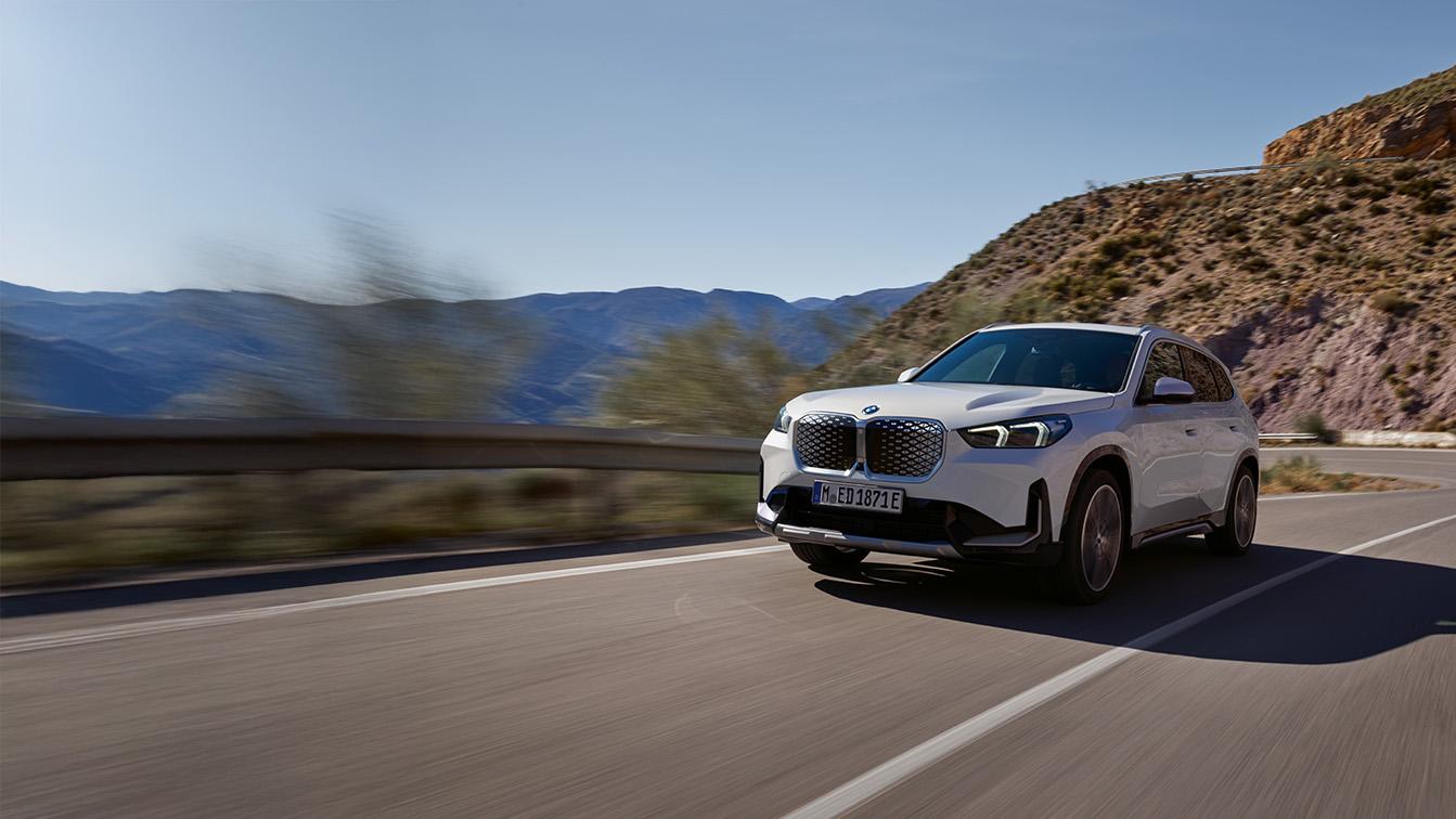 Ein weißer BMW iX1 fährt zügig auf einer kurvigen Bergstraße direkt auf den Betrachter zu. Während das Fahrzeug in der rechten Bildhälfte gestochen scharf abgebildet ist, erzeugen der verwischte Asphalt und die vorbeiziehende Leitplanke links eine starke Dynamik. Im Hintergrund erstrecken sich bläuliche Gebirgszüge unter einem klaren Himmel, während rechts ein sonnenbeschienener, karger Hang aufragt. Die markante Front mit dem geschlossenen, hellen Kühlergrill und den präzisen LED-Scheinwerfern verleiht dem Elektro-SUV eine moderne und kraftvolle Präsenz.