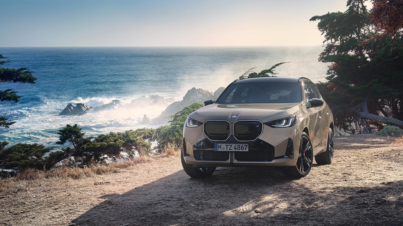 Das Bild zeigt den neuen BMW X3 in einem sandfarbenen Goldton, der auf einer Anhöhe direkt an einer rauen Küste parkt. Im Hintergrund bricht sich das Meer an schroffen Felsen, während das warme Sonnenlicht eine abenteuerliche und zugleich luxuriöse Atmosphäre schafft. Mit seiner markanten, großen Doppelniere und den dunklen Akzenten wirkt das SUV in dieser naturbelassenen Umgebung besonders robust und präsent.
