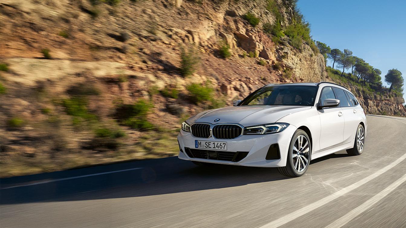 Das Bild zeigt einen weißen BMW 3er Touring (Kombi), der in Bewegung auf einer kurvigen Bergstraße aufgenommen wurde. Das Auto fährt von links nach rechts auf den Betrachter zu und ist im Halbprofil zu sehen. Im Hintergrund erhebt sich eine steile, felsige Böschung mit spärlicher Vegetation und einigen Bäumen am oberen Rand. Die Straße hat zwei Fahrspuren mit weißen Markierungen. Die Lichtverhältnisse sind hell und die Szene wirkt dynamisch, als würde das Auto gerade eine Kurve durchfahren.