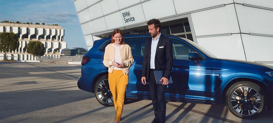 Ein Mann im Anzug mit einem Tablet und eine Frau in gelber Hose und beigefarbener Jacke mit einem Smartphone gehen lächelnd auf dem Parkplatz vor einem modernen BMW Servicegebäude. Ein blauer BMW SUV ist im Hintergrund geparkt.