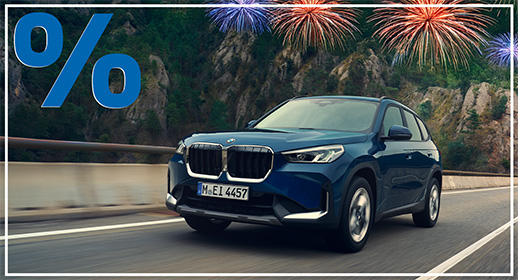 Dieses Bild zeigt einen dunkelblauen BMW X1, der zügig auf einer asphaltierten Straße entlang einer imposanten Bergkulisse fährt. Das kompakte SUV ist in einer dynamischen Schrägansicht von vorne aufgenommen, wobei die bewaldeten Felsen im Hintergrund dem Motiv eine abenteuerliche Atmosphäre verleihen. Über den Berggipfeln erstrahlen mehrere farbenfrohe Feuerwerke in Blau- und Orangetönen am Himmel. Wie bei den anderen Bildern der Serie wird die Grafik durch ein großes blaues Prozentzeichen in der oberen linken Ecke und einen feinen weißen Rahmen vervollständigt.